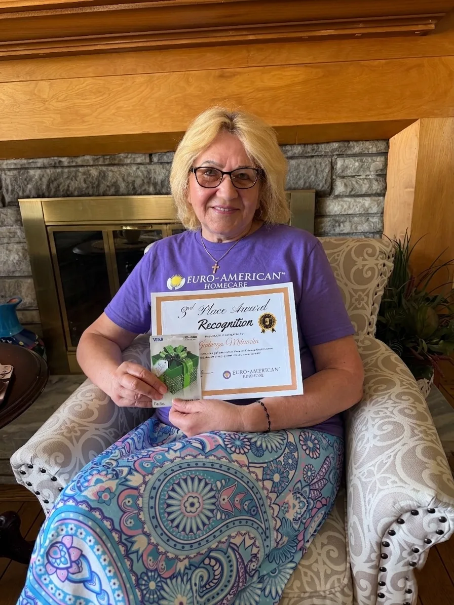 A caregiver seated by the fireplace with her award certificate and gift card